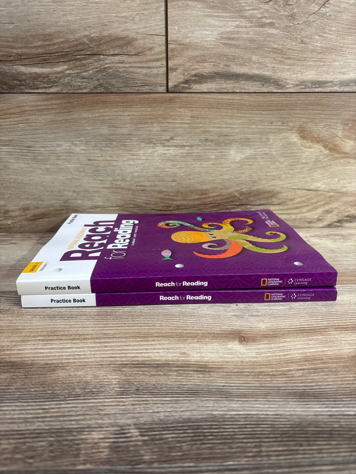 Two purple Reach for Reading Grade 2 practice books stacked on a wooden surface