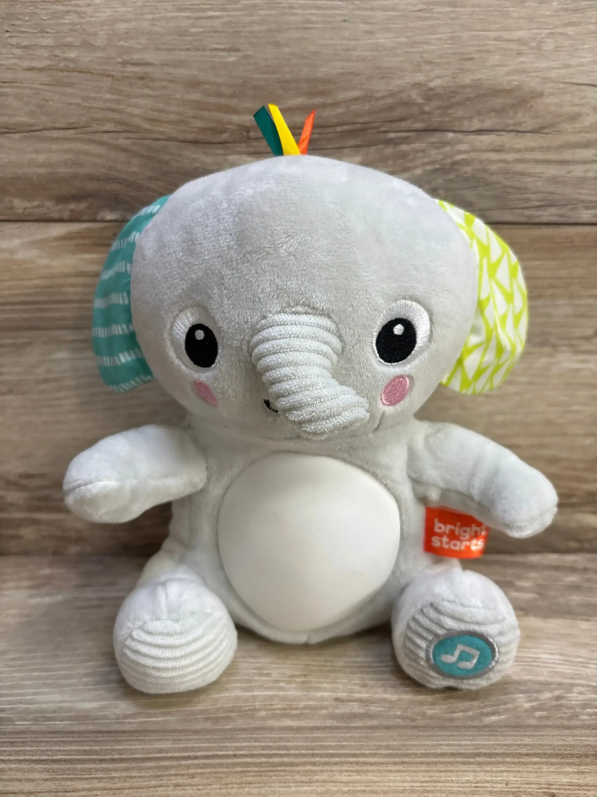 Front of Hug-a-Bye Baby Elephant plush with white belly, patterned ears, textured trunk and music button on wood background