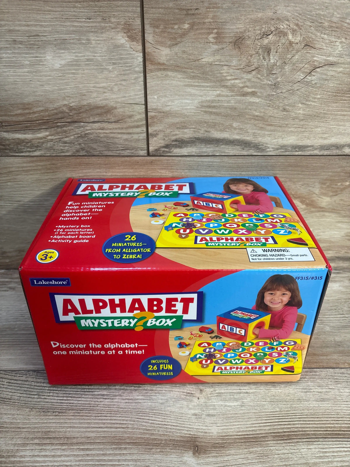 Front view of Lakeshore Alphabet Mystery Box with red packaging, alphabet board, and 26 miniature objects on wood flooring
