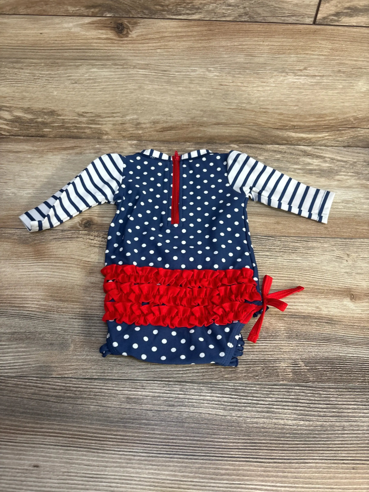 Flat lay of newborn navy polka dot long-sleeve rashguard swimsuit showcasing red ruffle tiers, striped sleeves, back zipper, and side bow on wood background