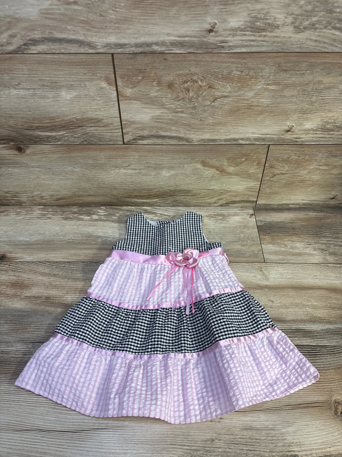 Flat-lay of toddler seersucker gingham dress in black-and-white and pink with ribbon flower at waist on wood floor, size 18M