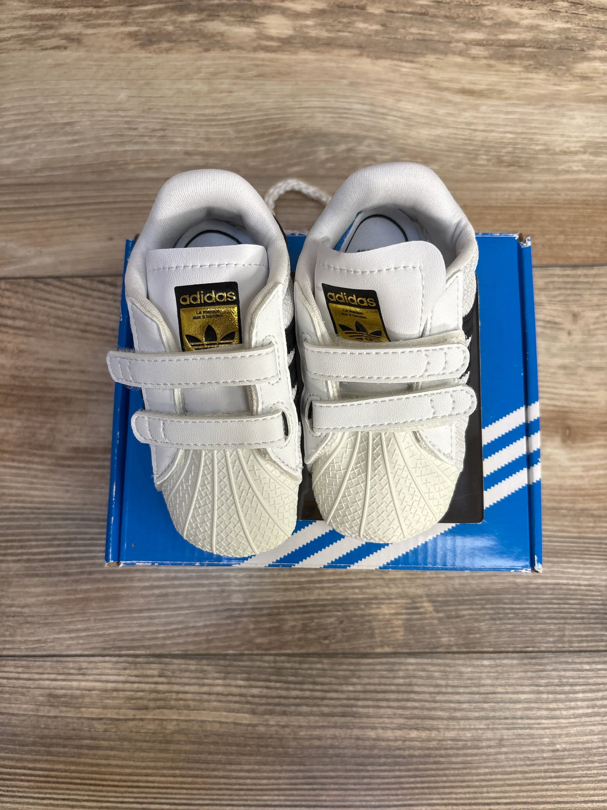 White Adidas infant sneakers with gold logo on box, wooden floor background