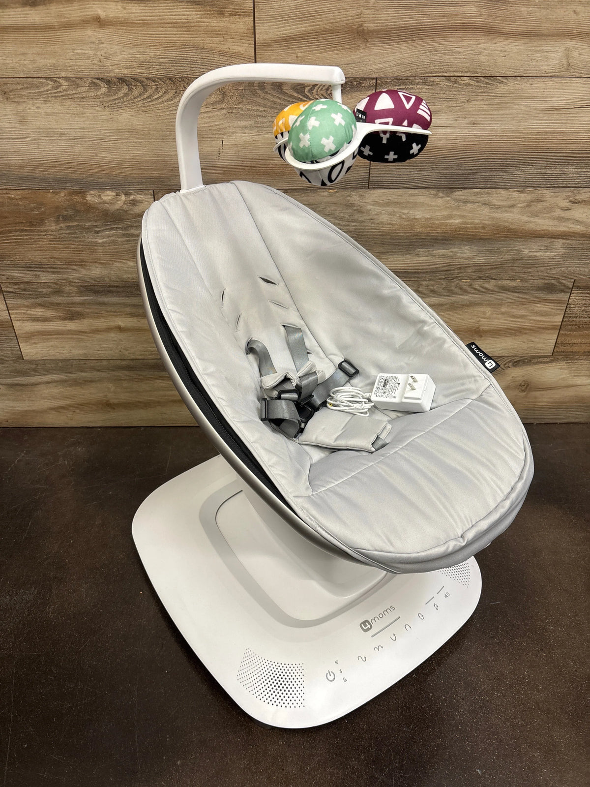 Three-quarter view of 4moms mamaRoo Multi-Motion Baby Swing with grey padded seat, harness, toy mobile on wood floor