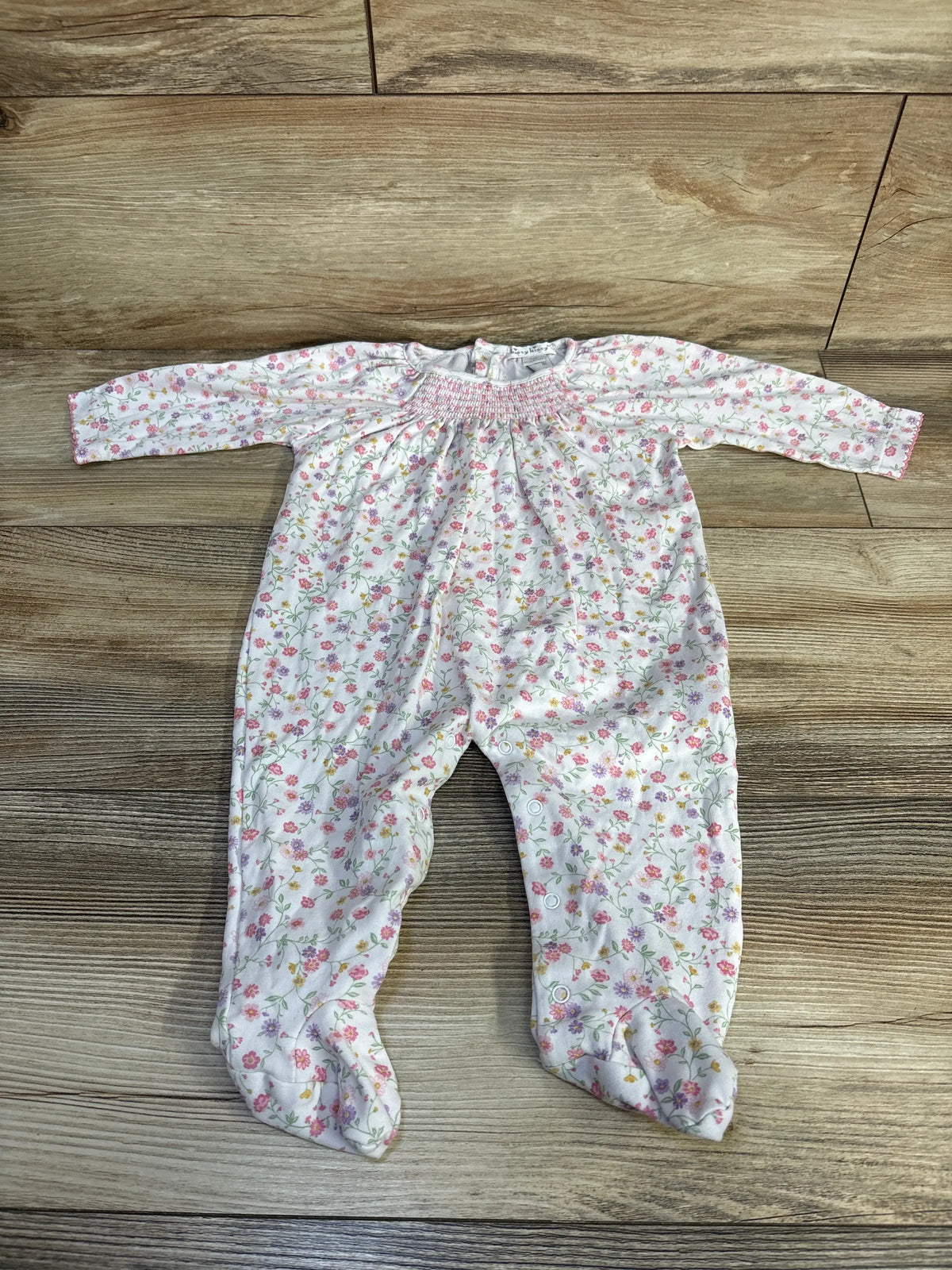 Top view of white Kissy Kissy floral coverall with long sleeves, smocked neckline, snap closures, and integrated feet on wood grain