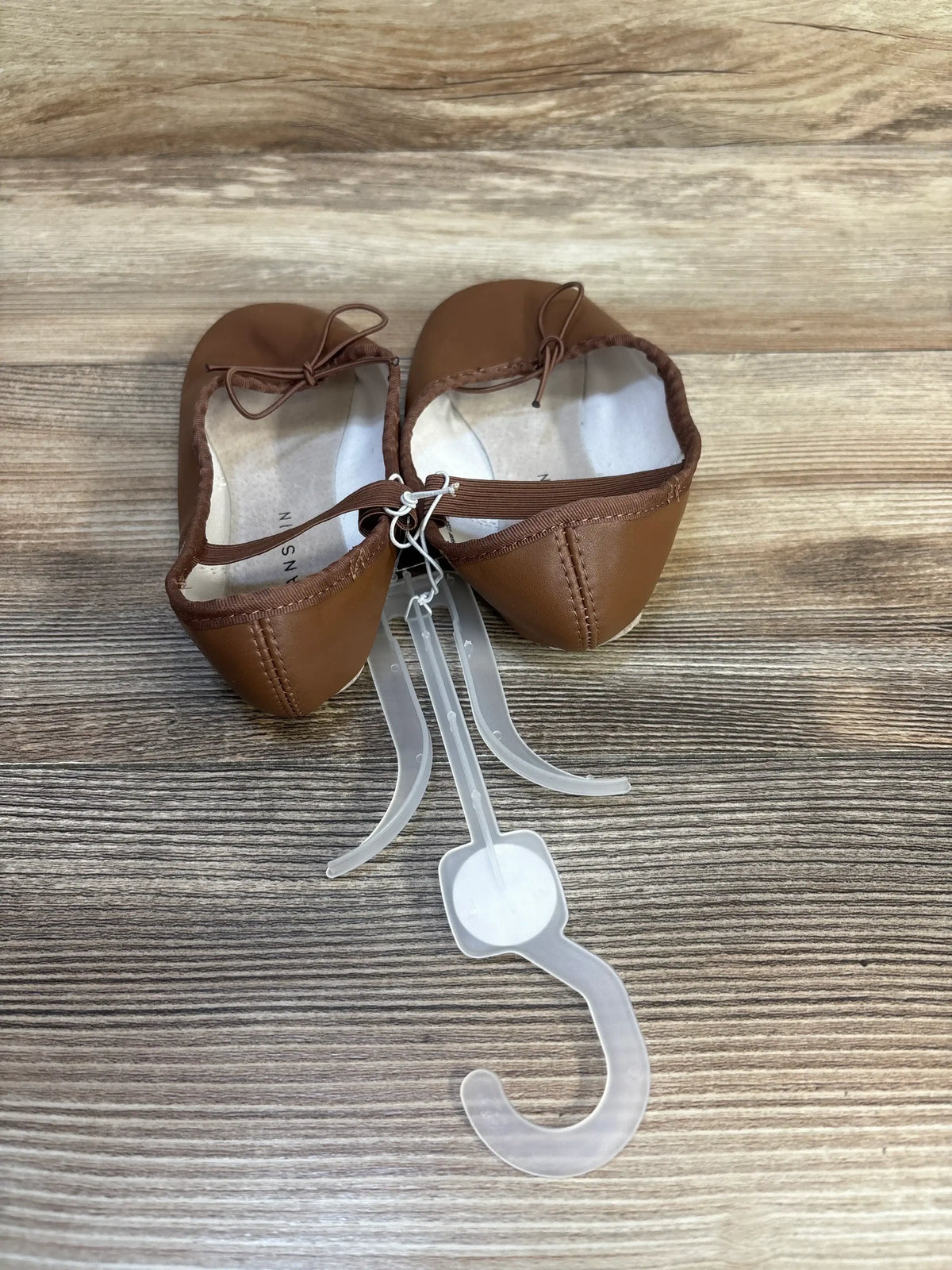 Top view of NEW Danskin Ballet Dance Flats in brown sz 13c showcasing bow accents and plastic display hanger with wood plank background