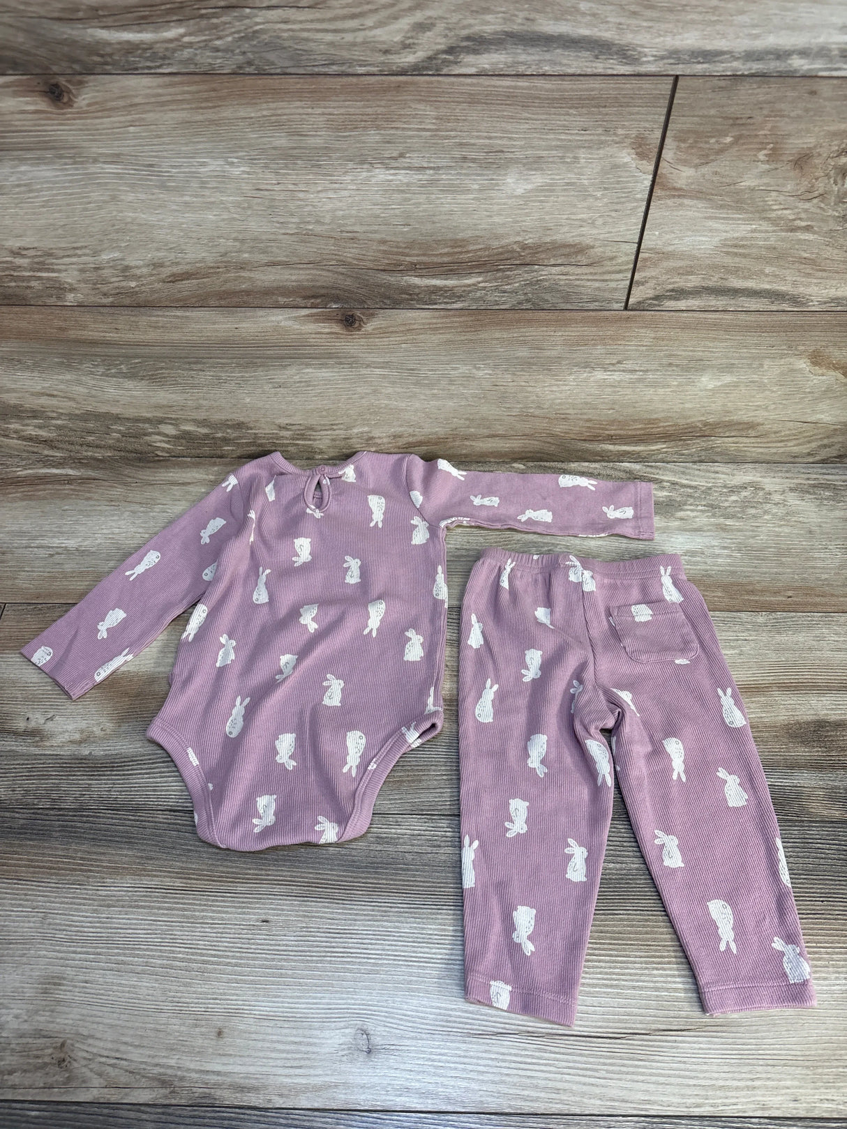 Pink baby onesie and pants set with white bunny print, soft ribbed fabric, laid on wood floor