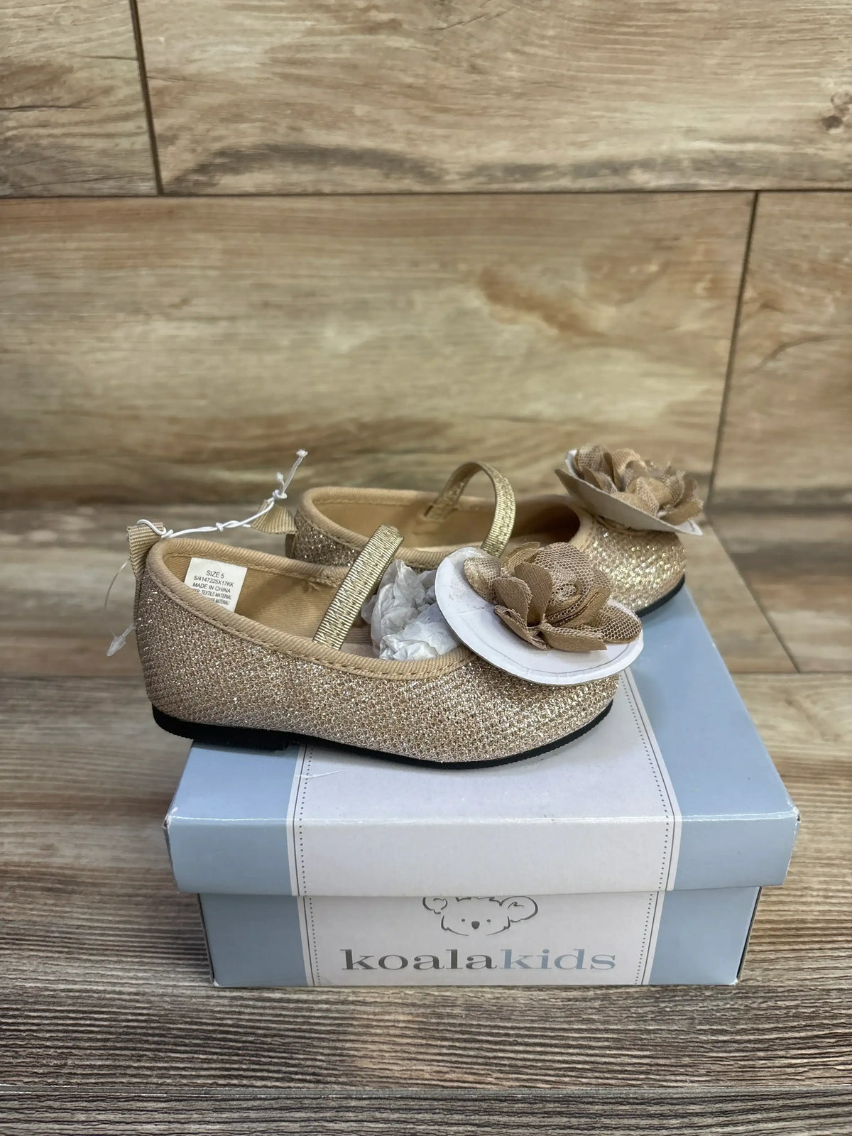 Side view of Koala Kids gold sparkle Mary Jane flats showcasing glitter finish and fabric flower embellishment on a shoebox