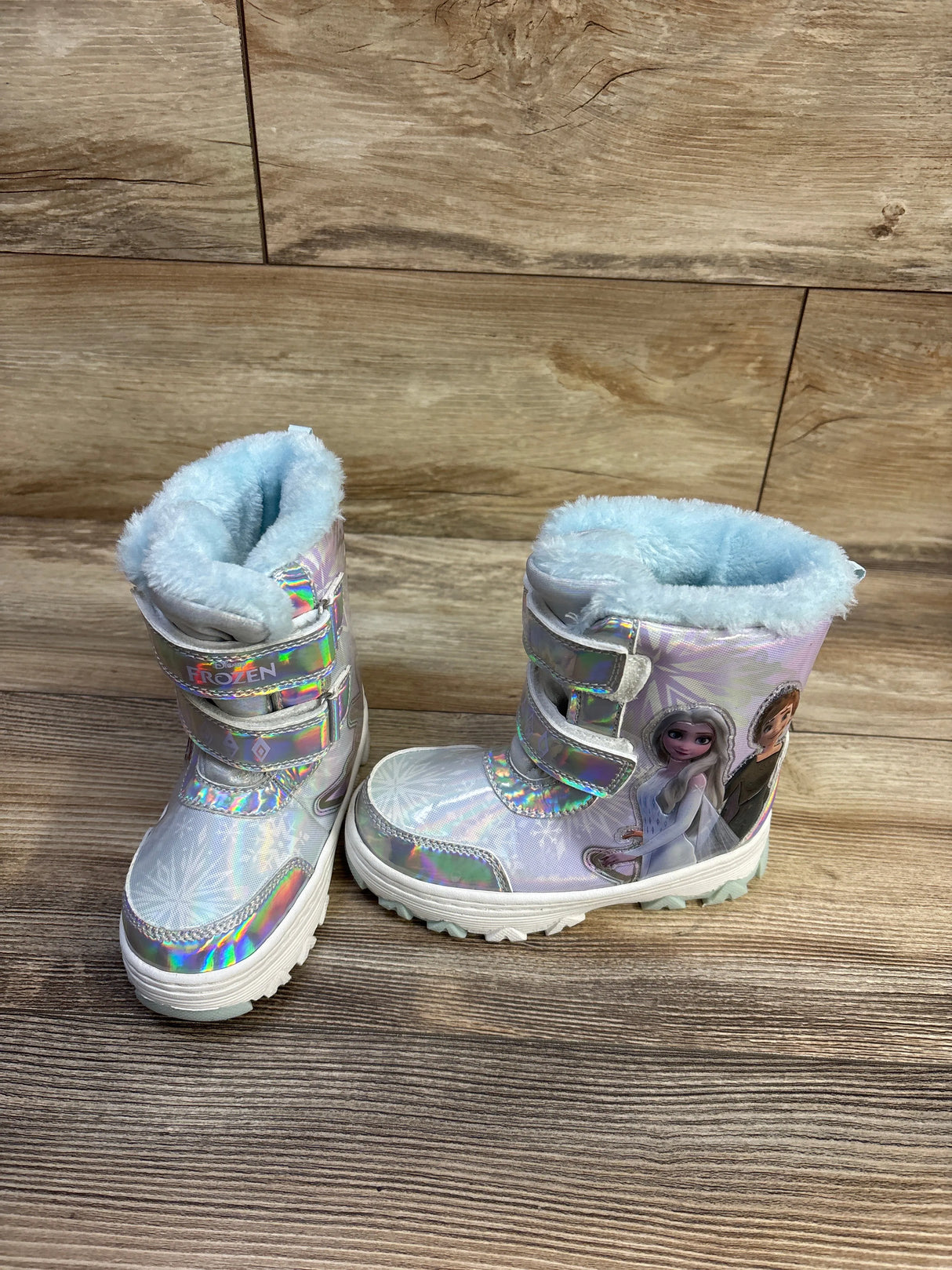 Three-quarter view of Disney Frozen silver holographic kids’ snow boots showcasing blue faux-fur lining, Velcro straps, and character graphic on a wood floor