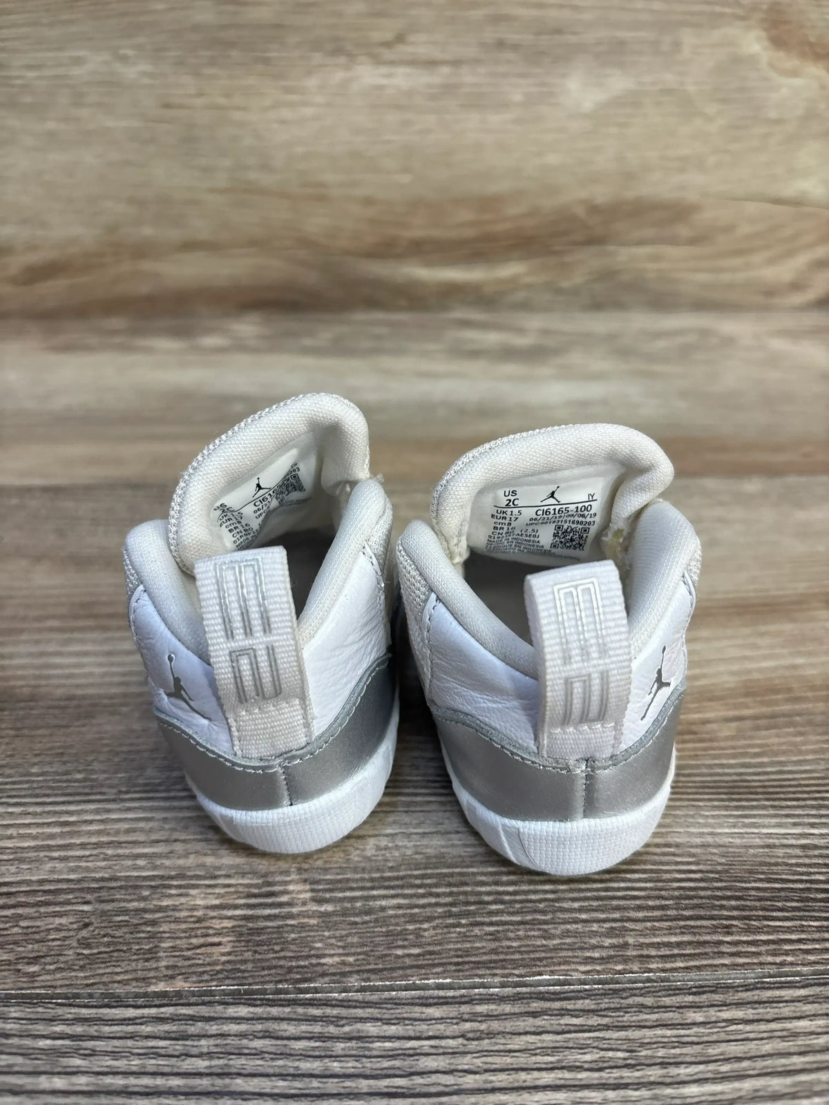 Back view of toddler Jordan sneakers showcasing white and silver leather pull tabs with size 2C label on wood plank floor
