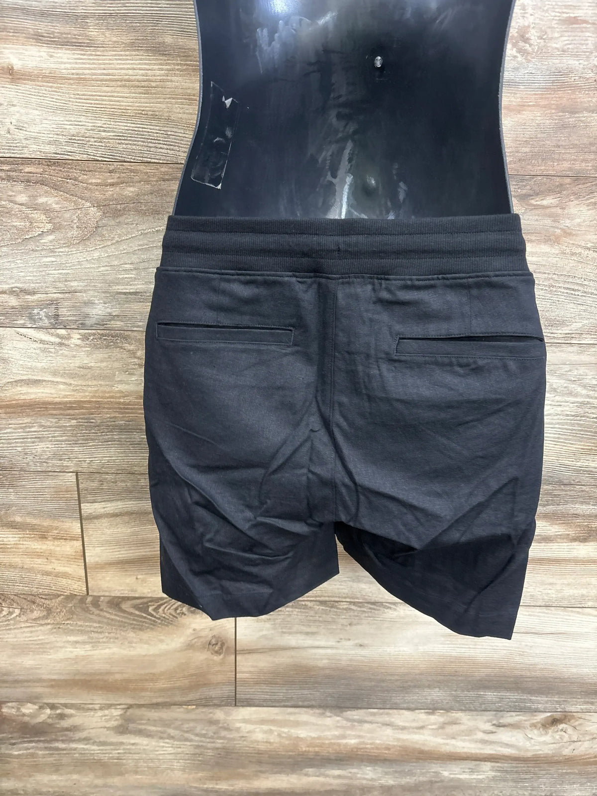 Back view of black maternity linen-blend shorts showcasing an under-belly elasticized waistband and back welt pockets on wood flooring