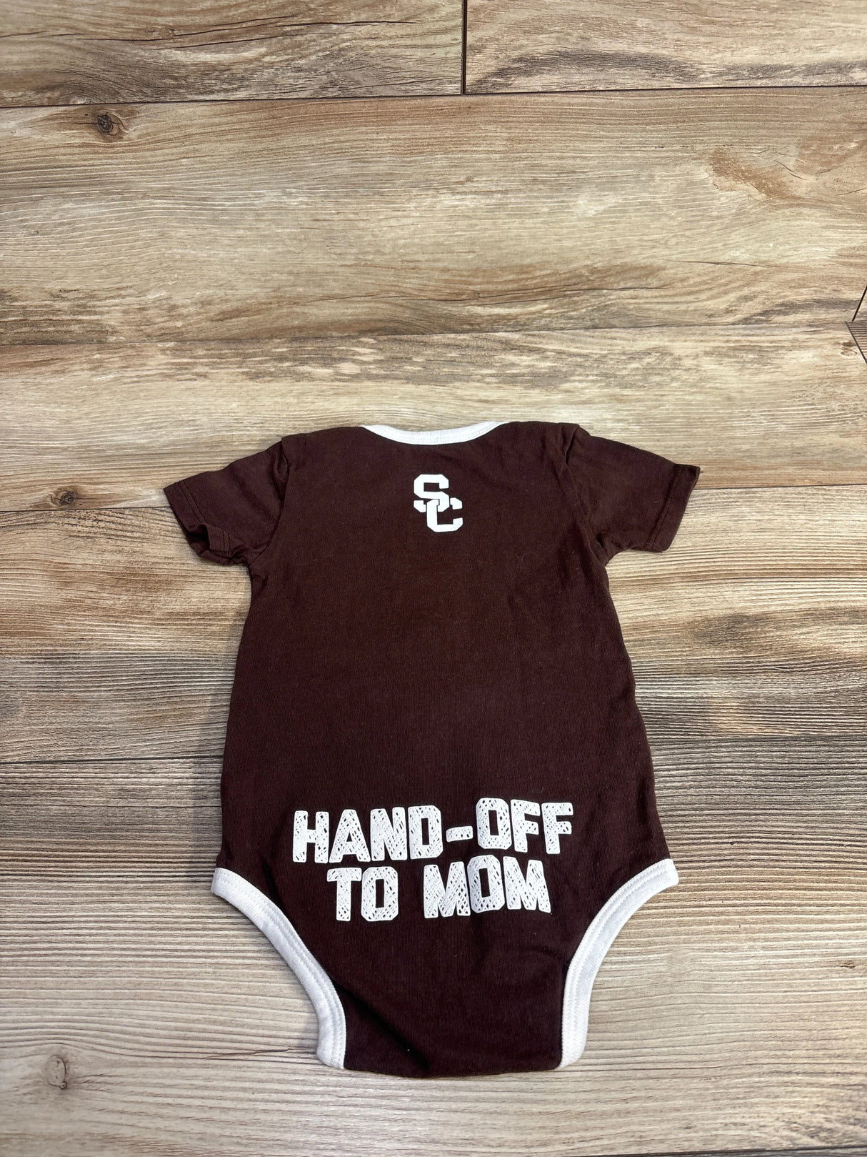 Brown baby onesie with white trim and text HAND-OFF TO MOM on back, displayed on wooden surface