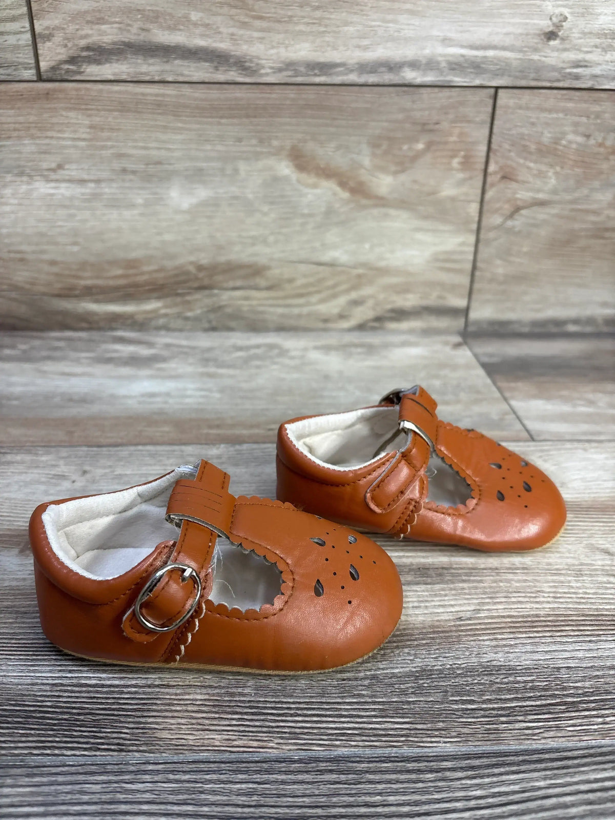 Front-side of Sienna chestnut baby Mary Jane sandals with scalloped edges, floral cutouts, buckle strap on light wood