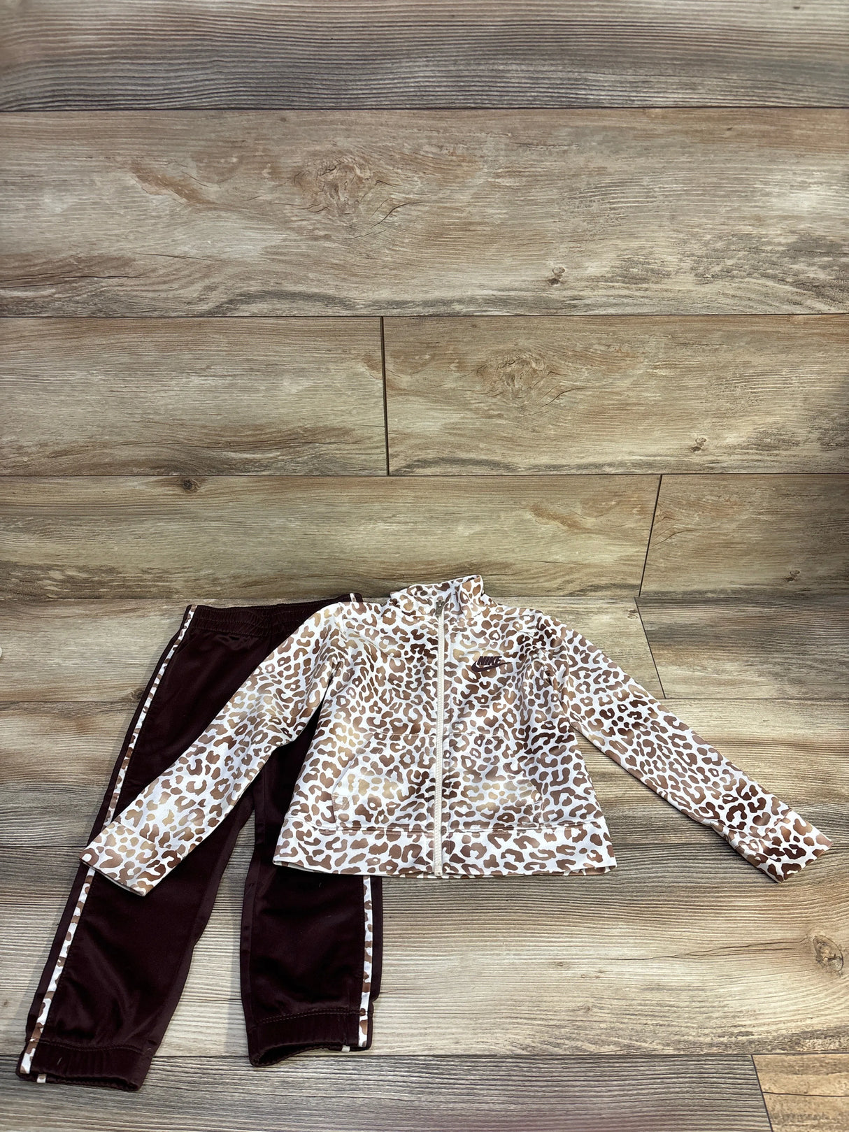 Flat lay of Nike toddler tracksuit, white leopard-print zip jacket and brown pants with leopard side stripes on wood floor
