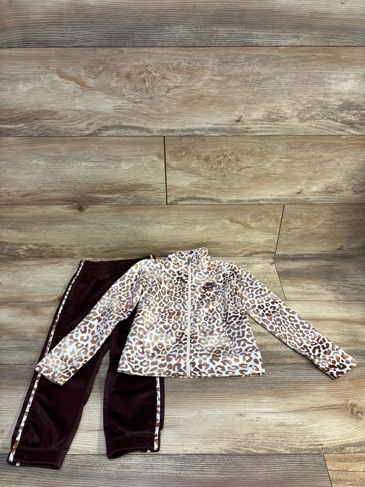 Flat lay of Nike toddler tracksuit, white leopard-print zip jacket and brown pants with leopard side stripes on wood floor