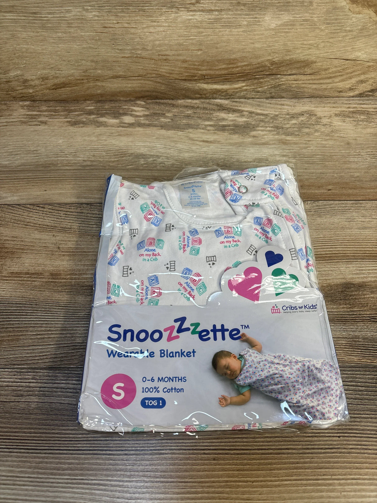 Front view of Snoozzzette wearable blanket (size S, 0–6 months, 100% cotton, TOG 1) in sealed packaging showing safe-sleep print on wood plank floor