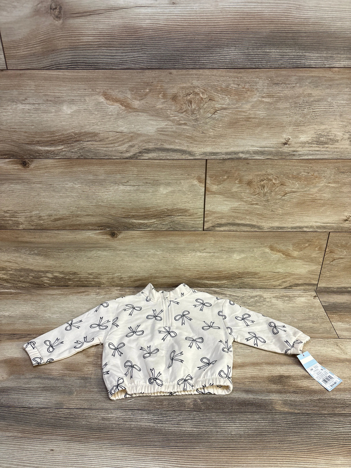 Flat lay of Cat & Jack quarter-zip ivory sweatshirt size 12M with black bow print, elastic hem and front zipper on wood