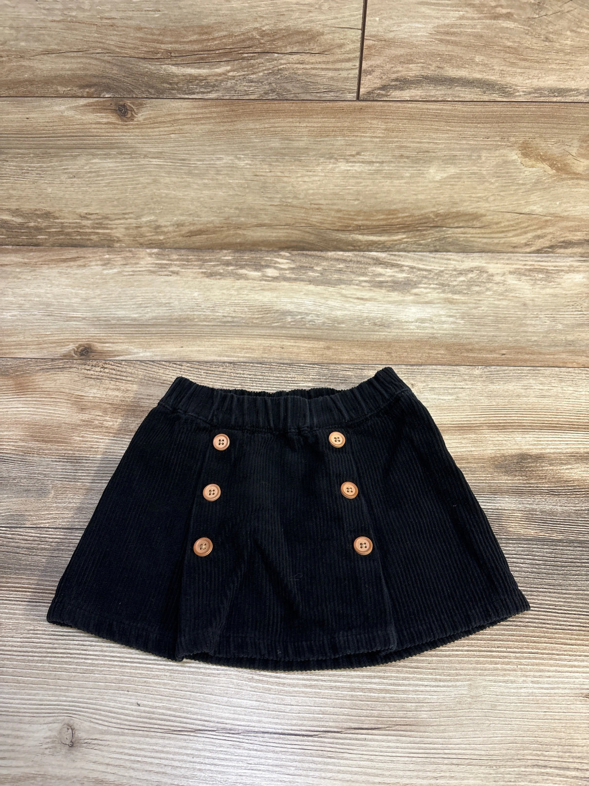 Top-down view of black corduroy toddler skirt showing two rows of wooden buttons and elastic waistband on light wood floor