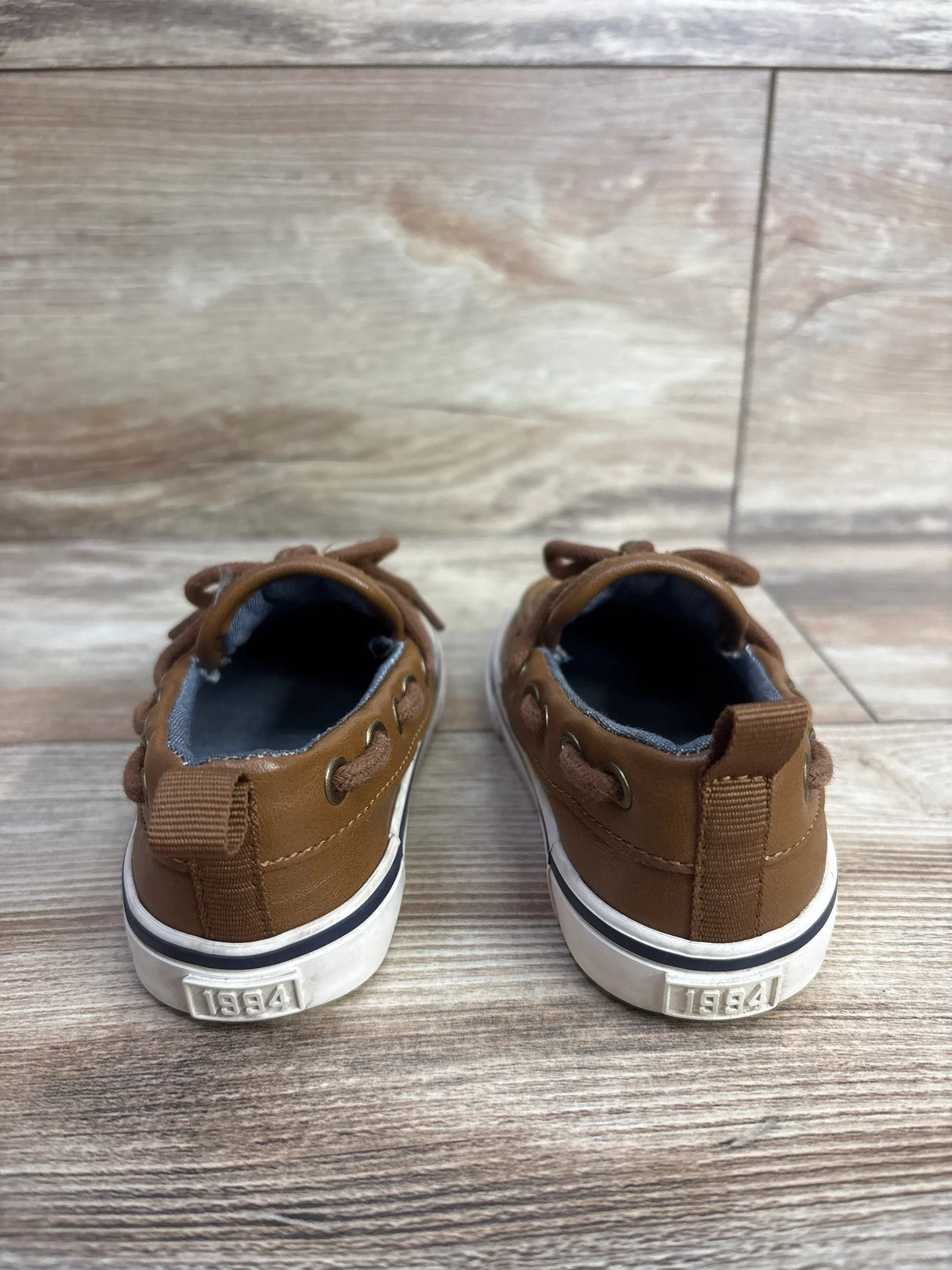 Pair of brown toddler shoes with white soles on wood floor, showing back view with 1994 detail