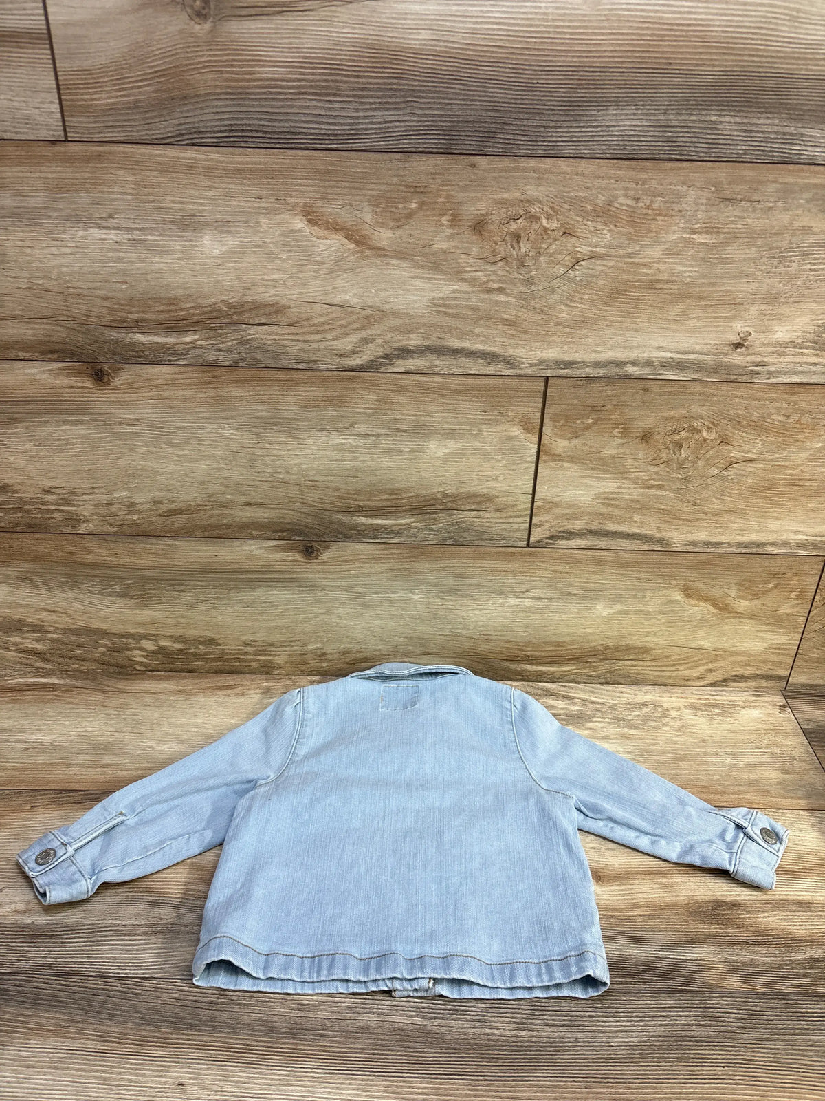 Back view of OshKosh blue denim jacket showcasing long sleeves and button cuffs on a wood floor