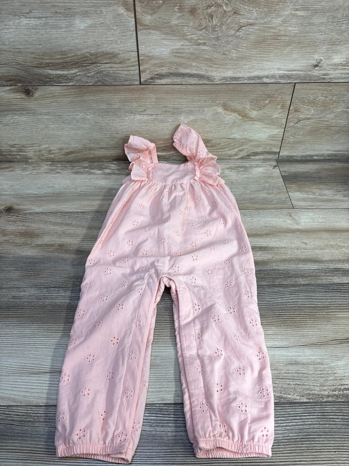 Front view of Carter’s pink eyelet sleeveless jumpsuit showcasing floral cutout details and ruffled straps on a wood-look floor