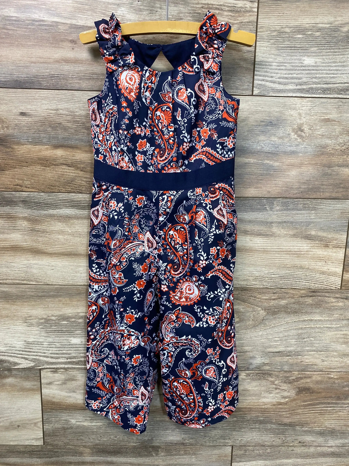 Front view of navy paisley toddler jumpsuit size 3T with ruffle shoulders and fitted waistband on wooden hanger against wood background