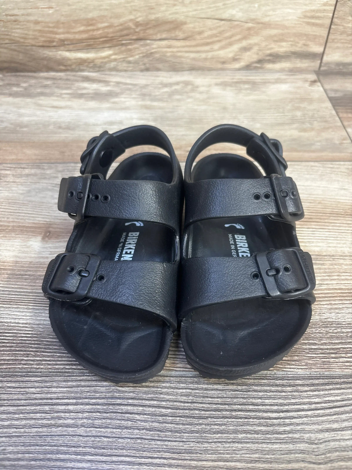Top view of black Birkenstock Milano EVA sandals showcasing three adjustable buckled straps and backstraps on a wood floor