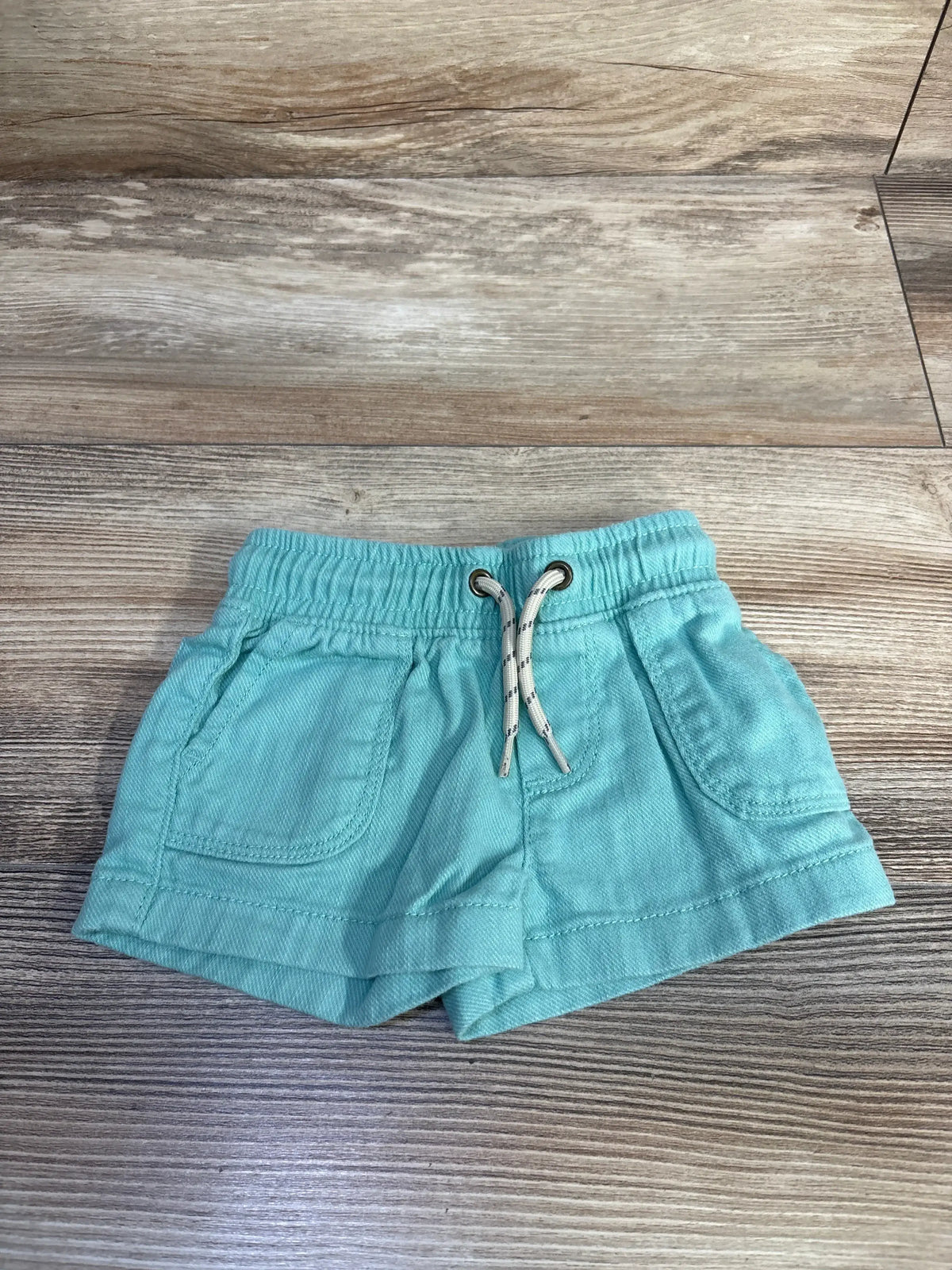 Flat lay of teal Carter's baby shorts showing elastic waistband with drawstring and front pockets on wood floor background