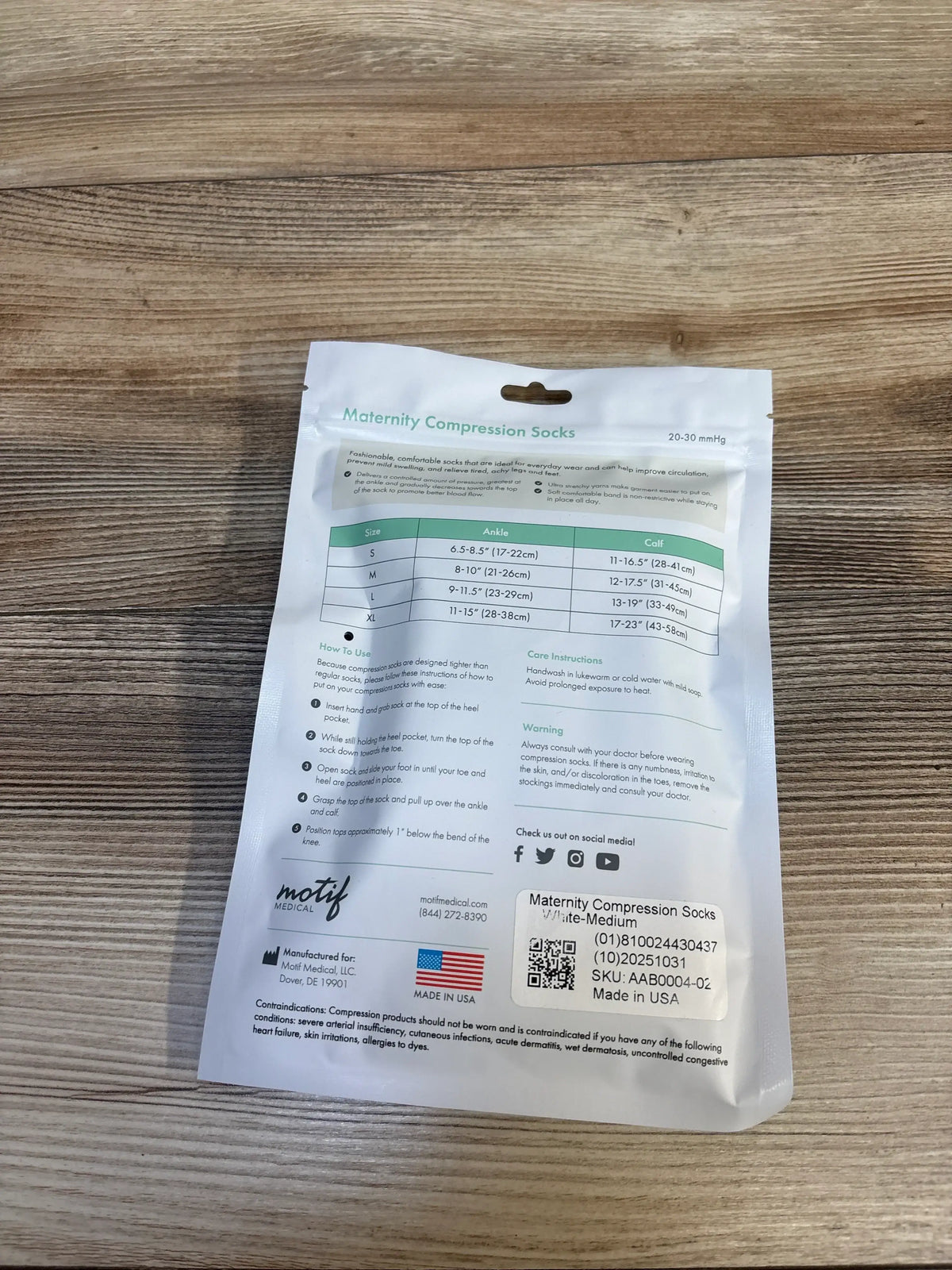 Back view of Motif Medical Maternity Compression Socks white sealed pouch with sizing chart and care instructions on wood plank floor