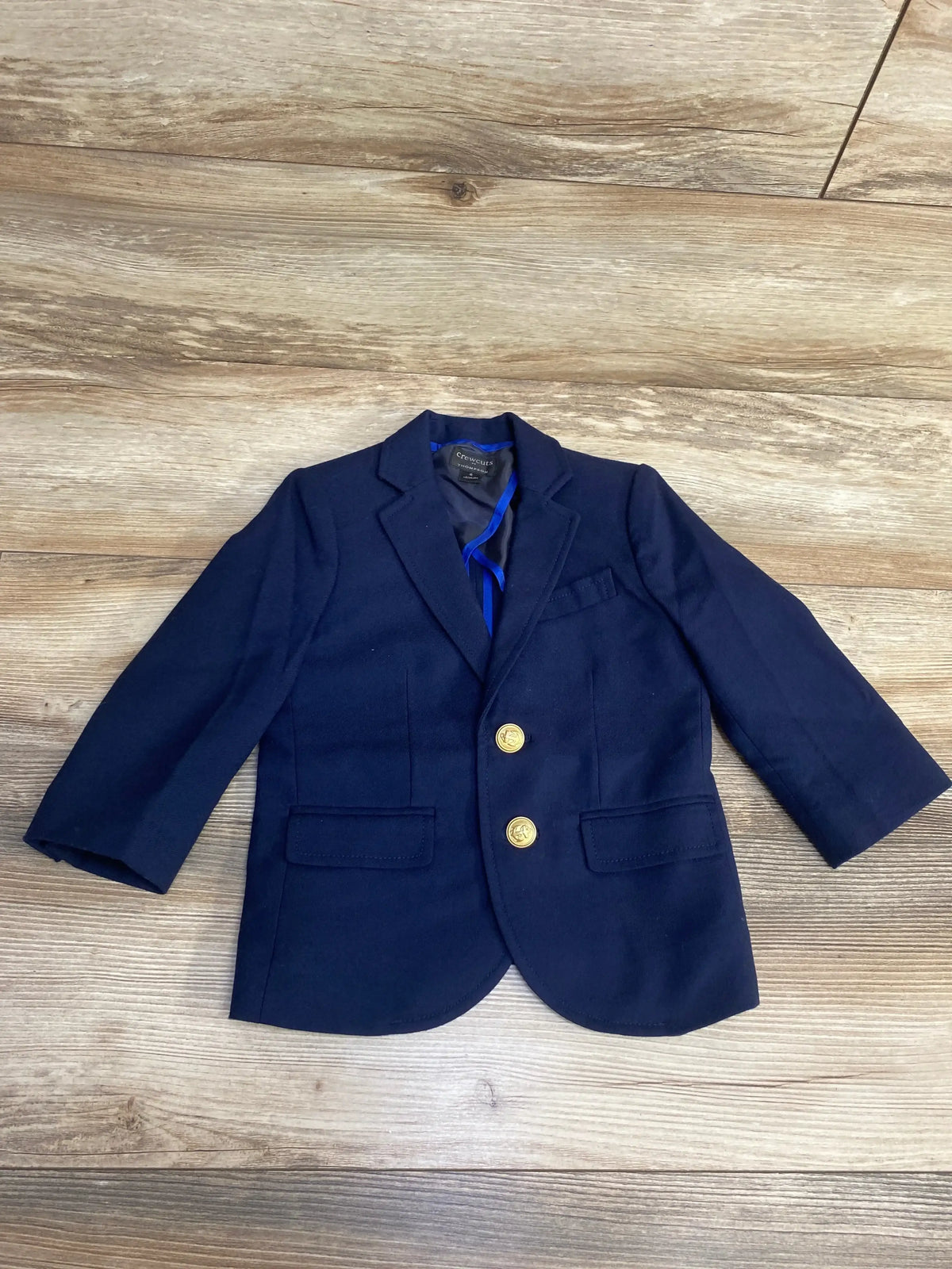 Front flat-lay of navy Crewcuts toddler blazer showing gold buttons, notch lapel and flap pockets on wood floor