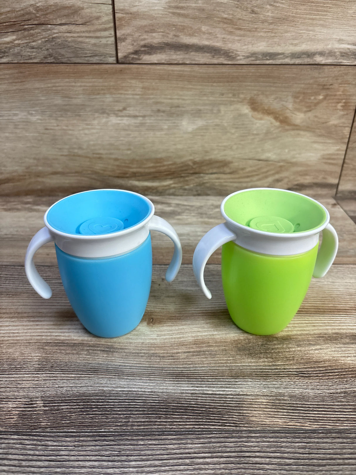 Front view of Munchkin Miracle 360 trainer cups two-pack in blue and green showcasing white handles and 360 degree spill-proof rim on wood background