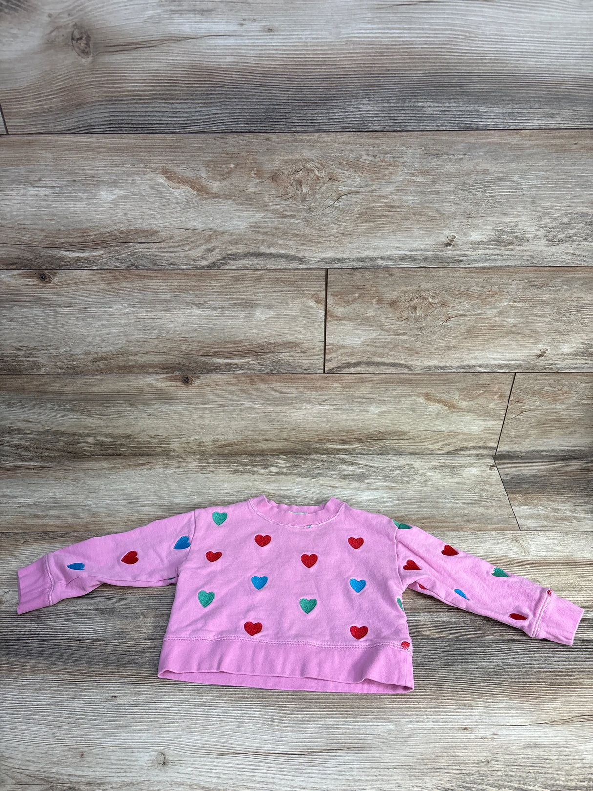 Pink children's sweatshirt with red, blue, and green heart patterns on a wood floor background