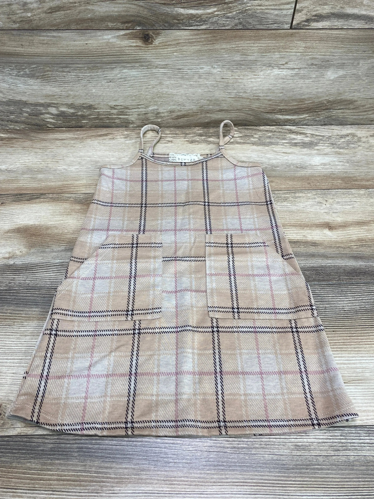 Top-down view of toddler brown plaid dress with two front patch pockets and spaghetti straps on a light wood background
