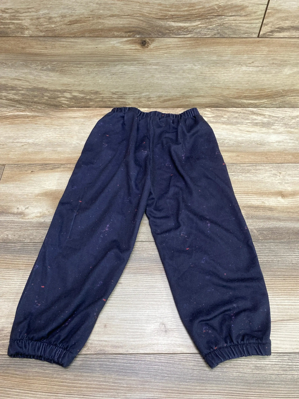 Top-down view of navy toddler joggers (size 2-3T) showing elastic waistband and elastic ankle cuffs with small pink specks on a light wood floor