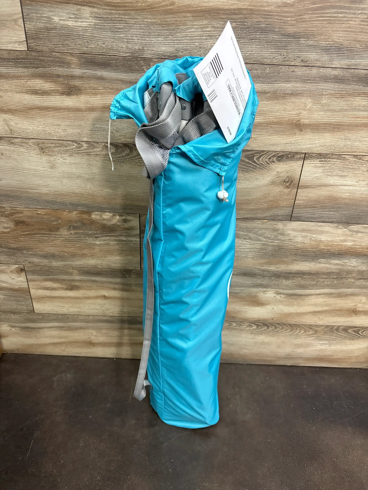 Folded view of Regalo My Play 6-panel play yard in teal carry bag with shoulder strap on wood flooring