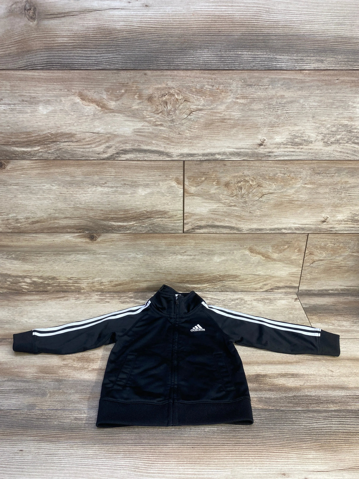 Front view of black Adidas toddler full-zip track jacket showing white three stripes on sleeves, chest logo, zip pockets on wood background