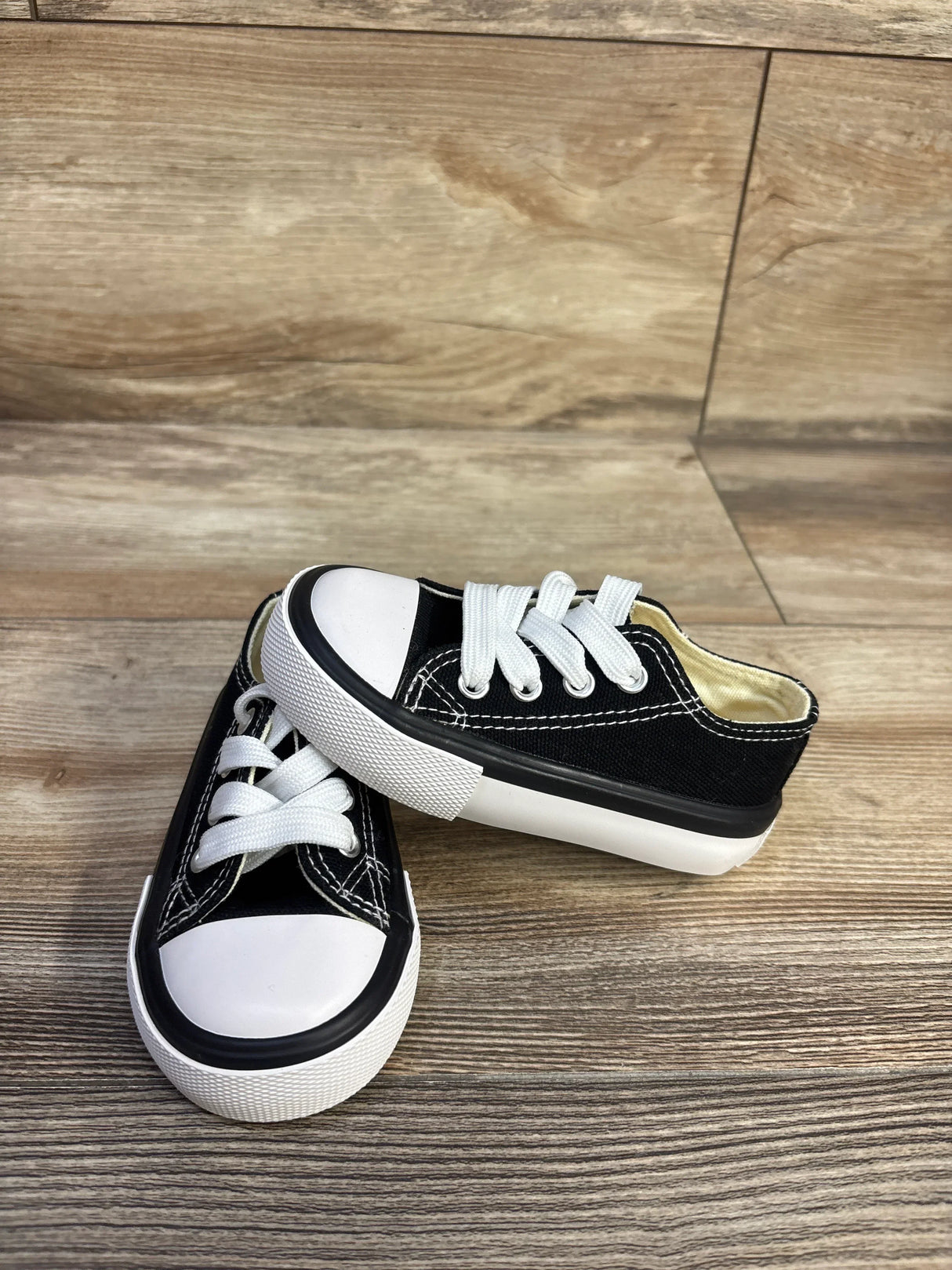 Three-quarter view of black low-top toddler sneakers with white rubber toe caps, white laces, and white soles on a wood floor background