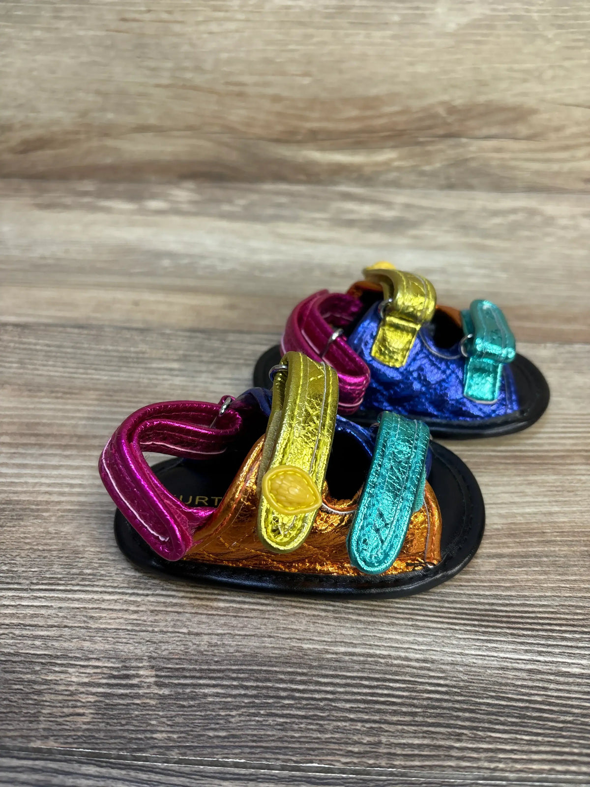 Front-left view of Kurt Geiger Orson metallic leather baby sandals size 2C showcasing multicolored Velcro straps on wood surface