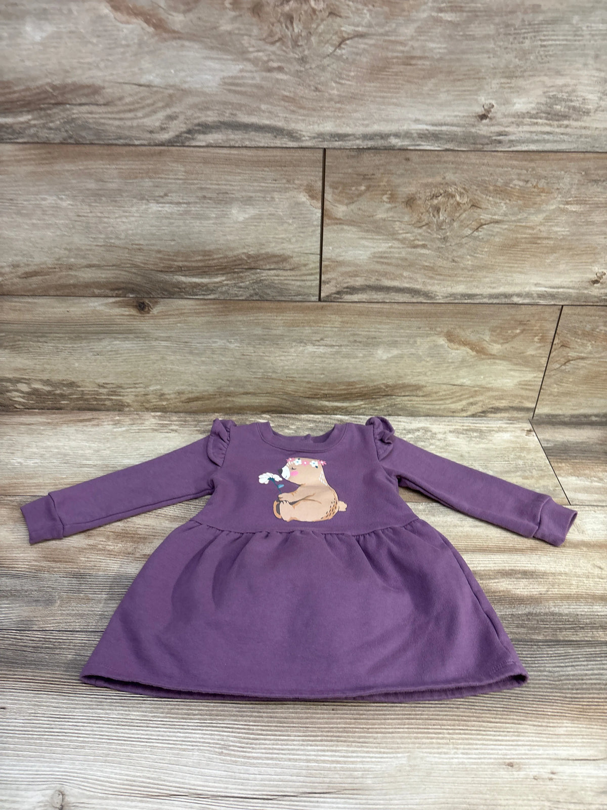Front view of purple baby sweatshirt dress showcasing long sleeves, ruffle shoulders, gathered skirt, and koala graphic on wood background