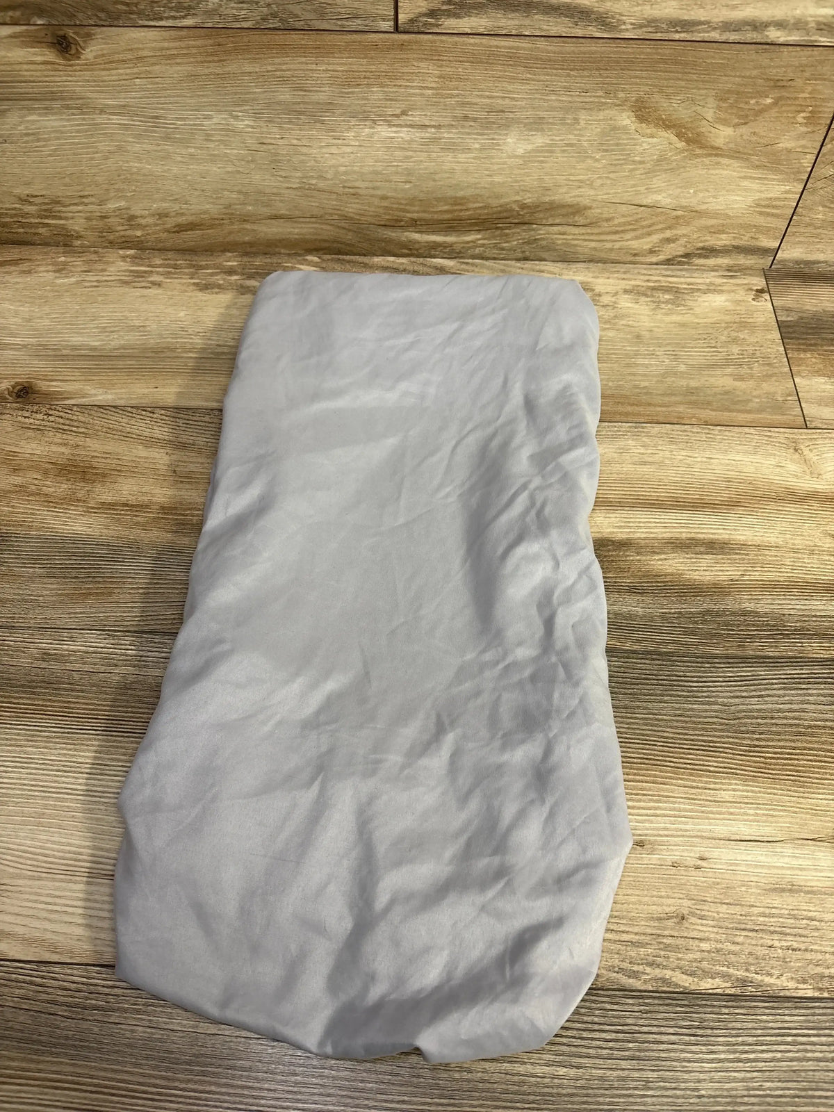 Top-down view of Parent’s Choice fitted bassinet sheet in light gray showing fitted shape and elastic edges on a wood floor background