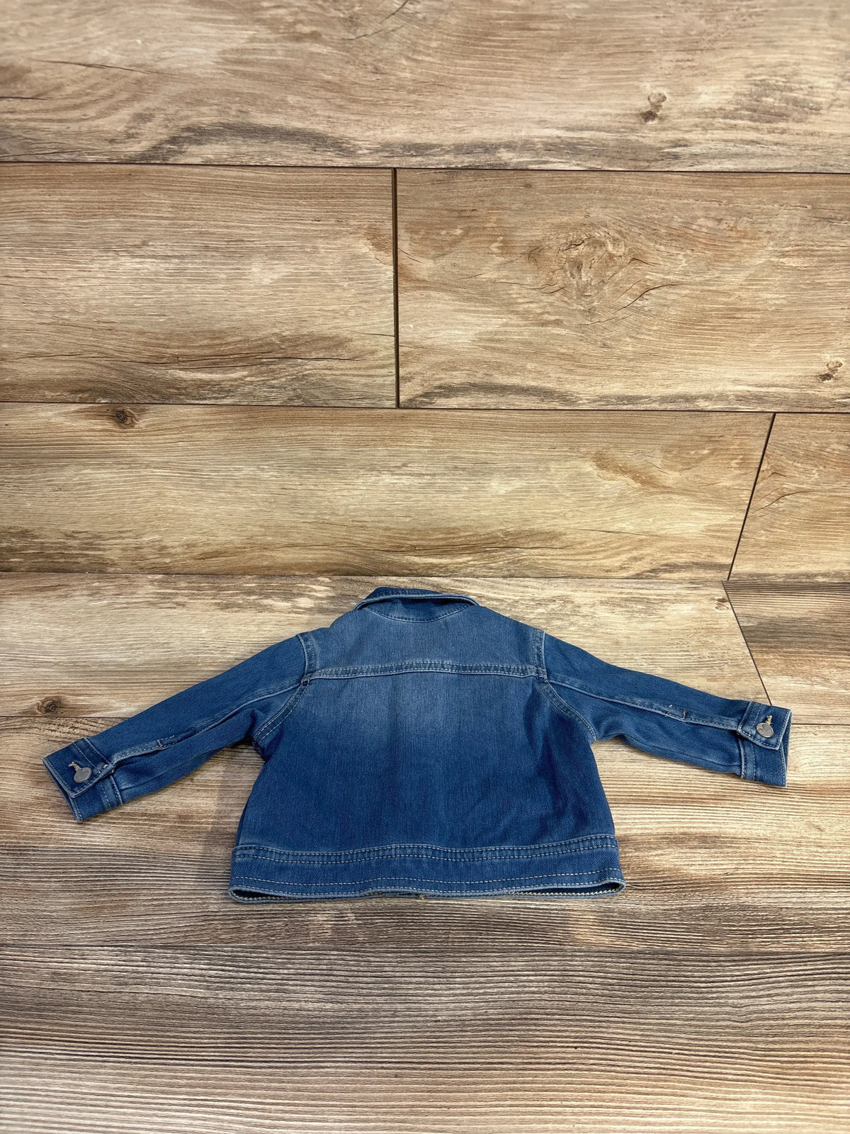 Back view of Old Navy blue denim jacket in size 3 to 6 months showing long sleeves and stitched seams on a wood floor
