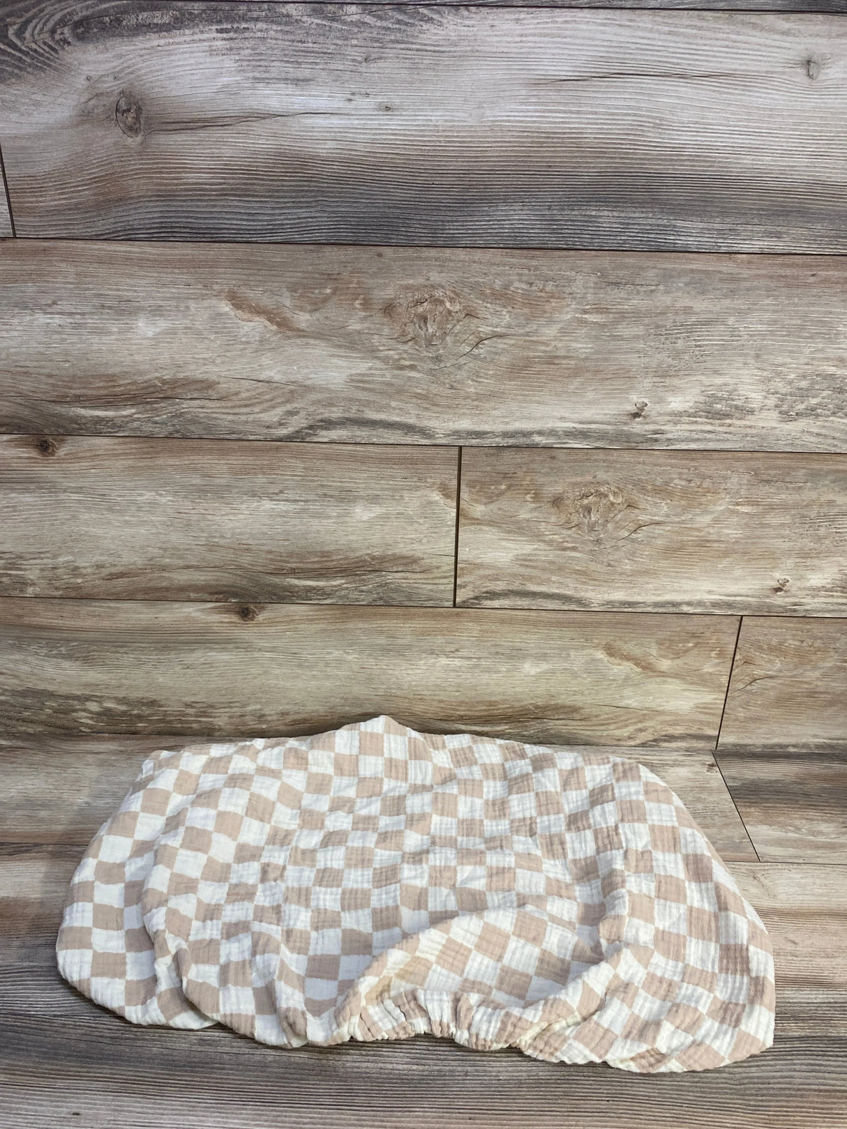 Flat lay of Brixton Phoenix muslin bassinet sheet showcasing checker pattern and elastic fitted edge on wood background