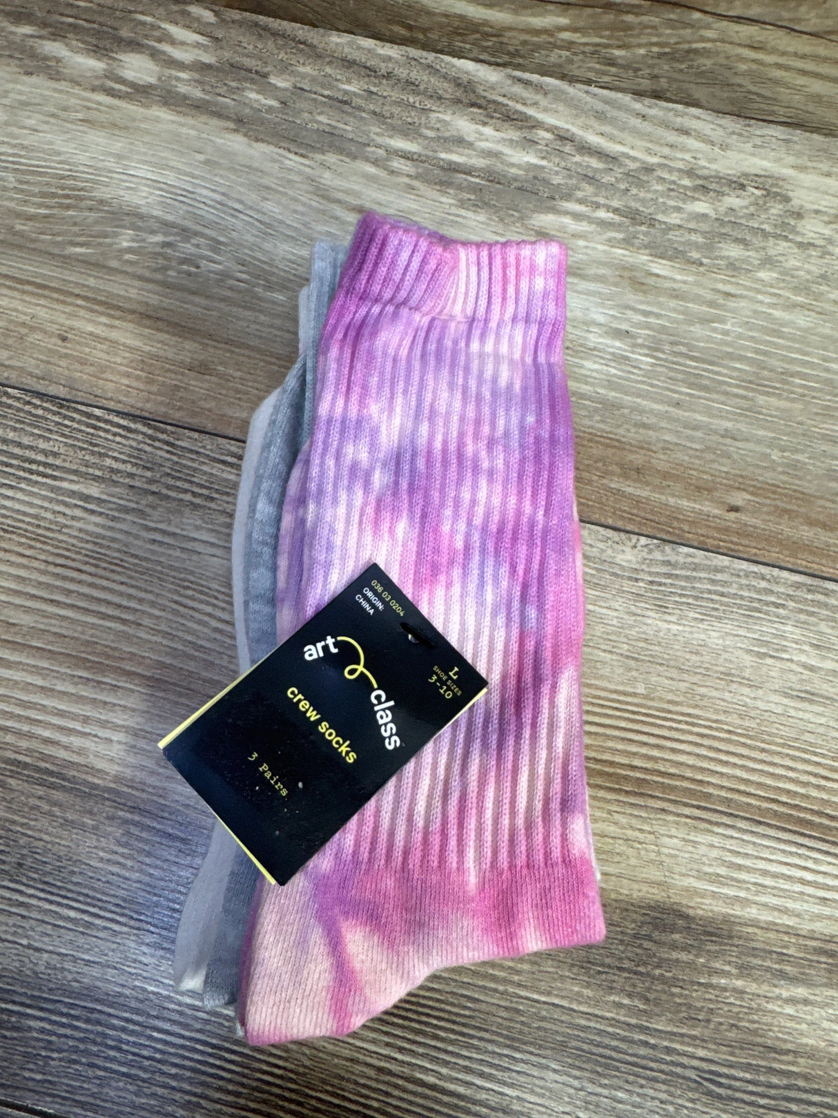 Top-down view of Art Class crew socks 3-pack showing pink tie-dye ribbed sock and gray pair with black product tag on wood floor