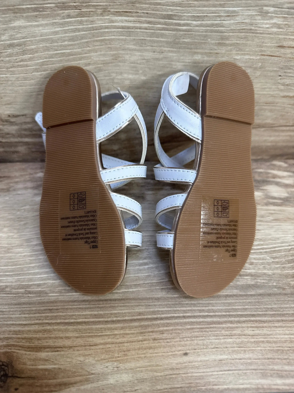 Bottom view of white Janie & Jack sandals showing brown rubber soles and metallic trim straps, size 7C on wood