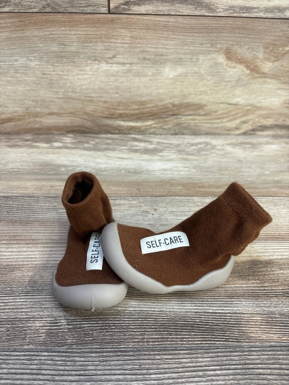 Side view of Tiny Explorings Self-Care Sock Shoes showcasing brown sock uppers and white soles on a wood-grain background