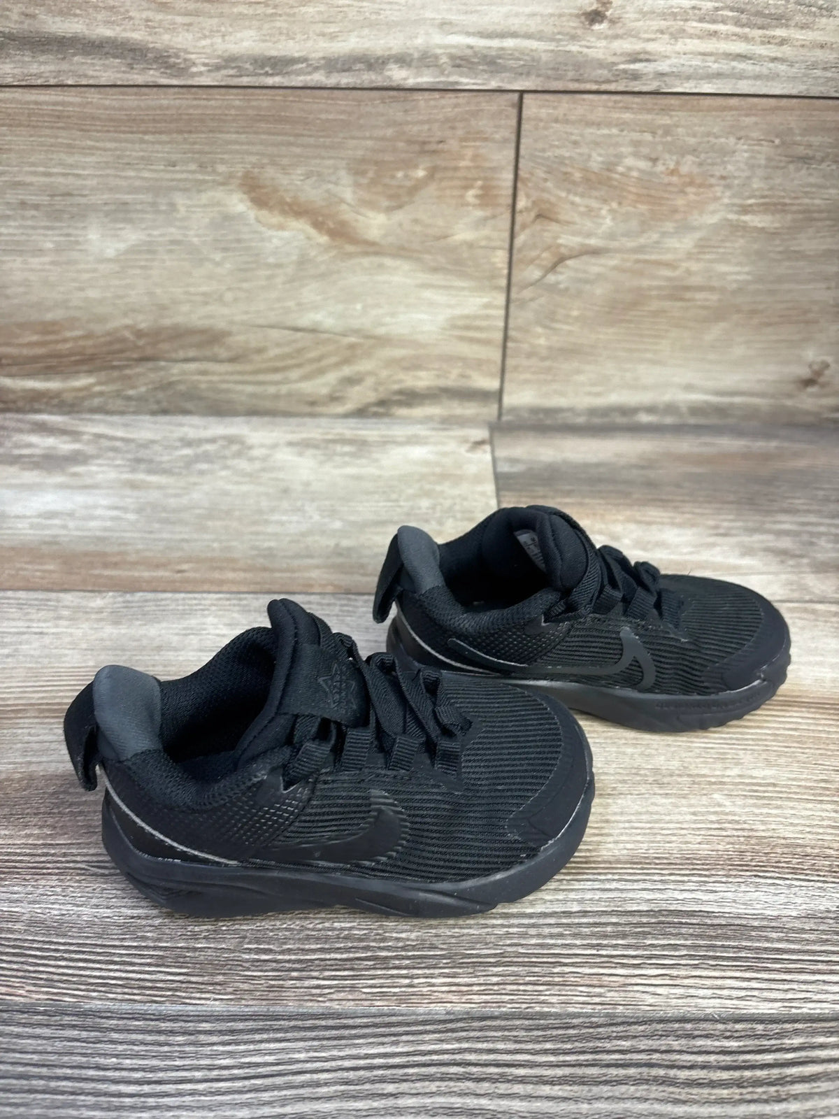 Front three-quarter view of toddler Nike Star Runner 4 sneakers showing black mesh upper, molded sole, heel pull tab and Nike swoosh on wood floor