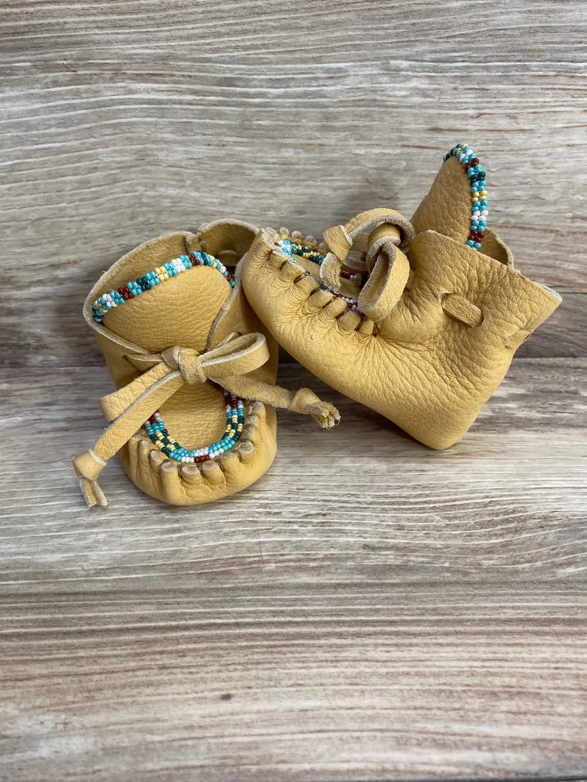 Front view of yellow leather baby moccasins showcasing hand-beaded multicolor trim and tied leather laces on wood background