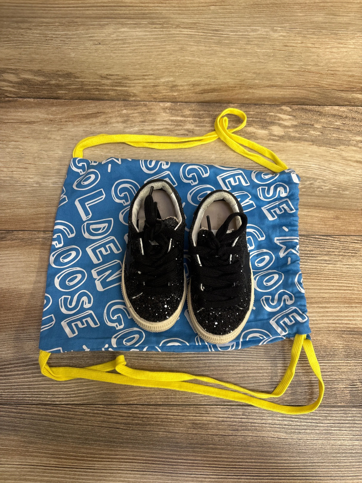 Black glitter toddler sneakers with white soles on a blue drawstring bag with yellow straps on wooden floor
