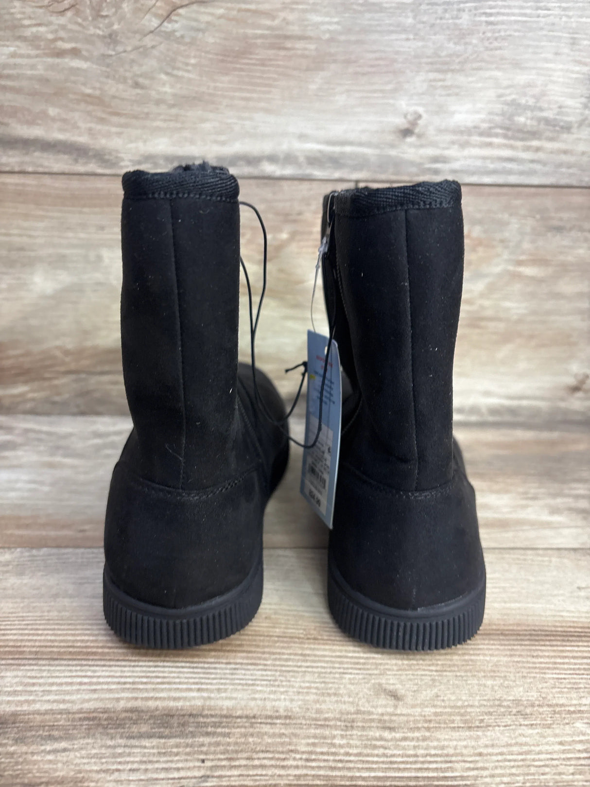 Back view of black kids suede winter boots showcasing rubber treaded soles with hangtag attached on wood plank floor