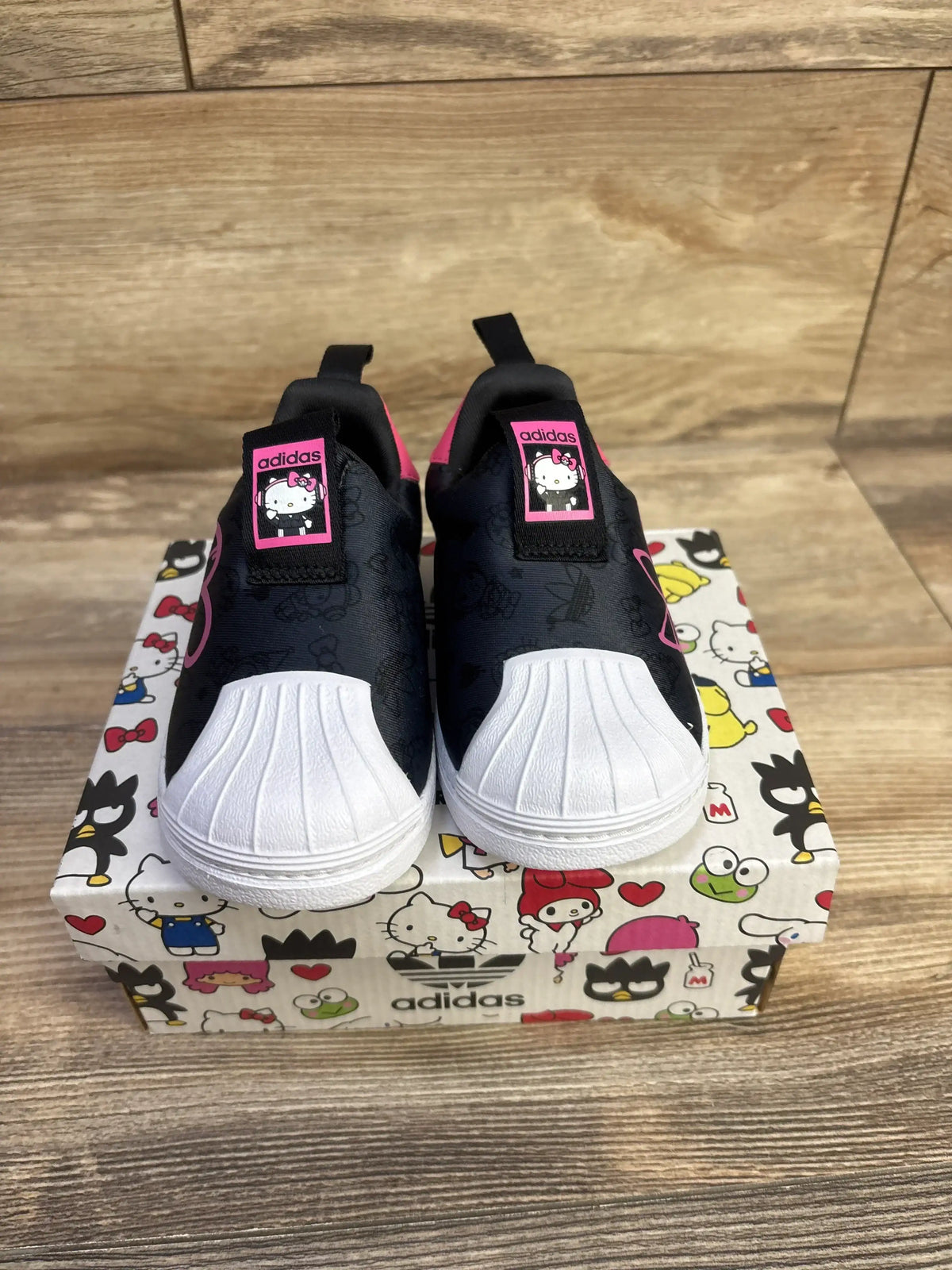 Front view of Adidas Hello Kitty Superstar 360 slip on sneakers in black showcasing white shell toe and pink Hello Kitty tongue patch on character print box