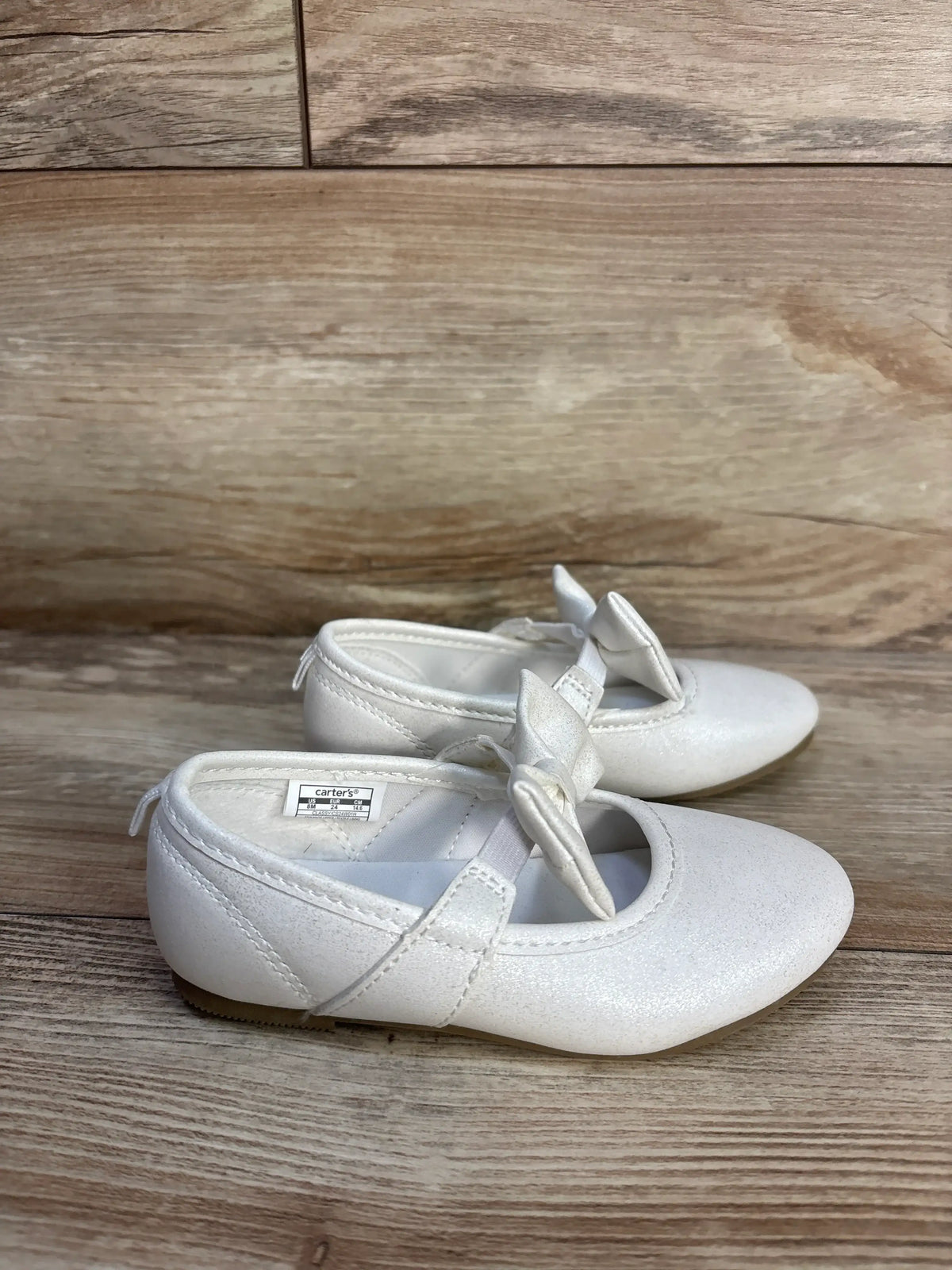 Pair of white shimmering Mary Jane flats with bows by Me n Mommy To Be on a wood surface.
